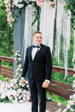 Siyah smokinli Avrupalı damat, arka planda düğün kemerinin önünde duruyor ve nikah töreninde genç karısını bekliyor. Aşk yeminleri ve alyans takası..