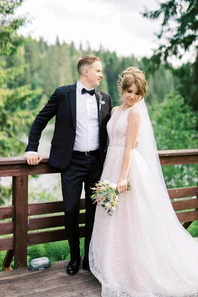 Full length portrait of handsome man in stylish black tuxedo suit embracing his charming young bride in white wedding dress. Happy newlywed couple posing near forest lake.