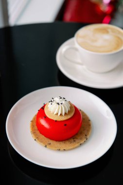 Kırmızı kremalı çilekli cheesecake keki. Siyah bir masada beyaz bir tabakta servis yapmak. Kadınlar için tatlılar günü. Kapuçinolu kahve fincanı..