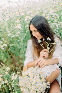 Beyaz elbiseli güzel esmer kadın papatya tarlasında baharın tadını çıkarıyor. Yaz kırsal konsepti. Doğa tatiline yakın. Kadın ve papatyalar. Yaz ışığı