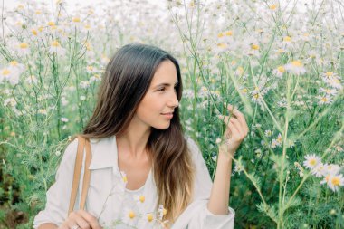 Beyaz elbiseli güzel esmer kadın papatya tarlasında baharın tadını çıkarıyor. Yaz kırsal konsepti. Doğa tatiline yakın. Kadın ve papatyalar. Yaz ışığı