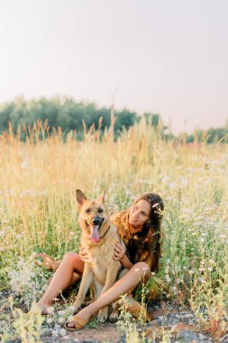 Esmer bir kız çayırda Alman çoban köpeğiyle yürüyor ve oynuyor. Arkaplanda gün batımı ve bulanık arkaplan.