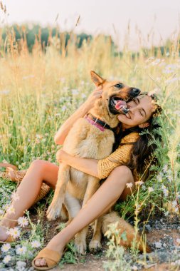 Bir Alman çoban köpeğini kucaklayan, güneşin altın ışınlarında çimlerin üzerinde oturan ve gün batımını izleyen bir kız. Kadına odaklan.