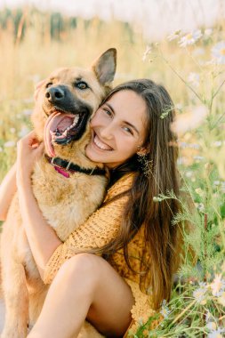 Esmer bir kız çimlerin üzerinde oturuyor ve bir Alman çoban köpeğiyle çayırda sarılıyor. Arka planda gün batımı ve bulanık arkaplan. Tatilde doğa yürüyüşü kavramı. Kahkaha ve duygular. Köpek insanı yalıyor..