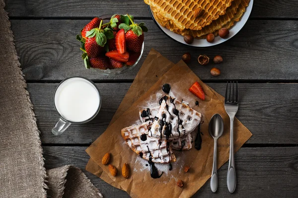 Gofres belgas caseros servidos sobre papel de hornear con salsa de chocolate, fresa, avellanas, almendras, azúcar en polvo y vaso de leche Imagen De Stock