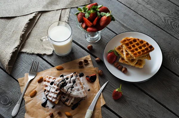 Gofres belgas caseros servidos sobre papel de hornear con salsa de chocolate, fresa, avellanas, almendras, azúcar en polvo y vaso de leche Fotos De Stock Sin Royalties Gratis