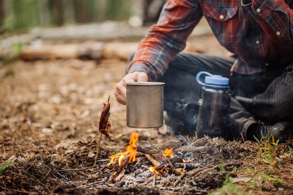 Чоловік мандрівник руки тримає кухоль з водою біля вогню на відкритому повітрі — стокове фото