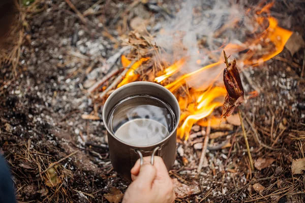 Чоловік мандрівник руки тримає кухоль з водою біля вогню на відкритому повітрі — стокове фото