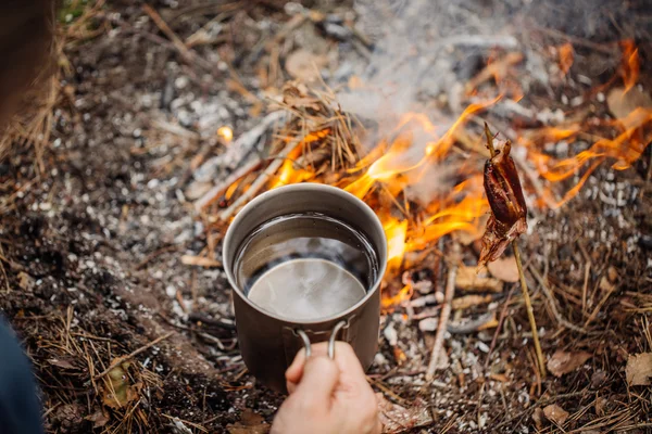 Homem viajante mãos segurando caneca com água perto do fogo ao ar livre — Fotografia de Stock