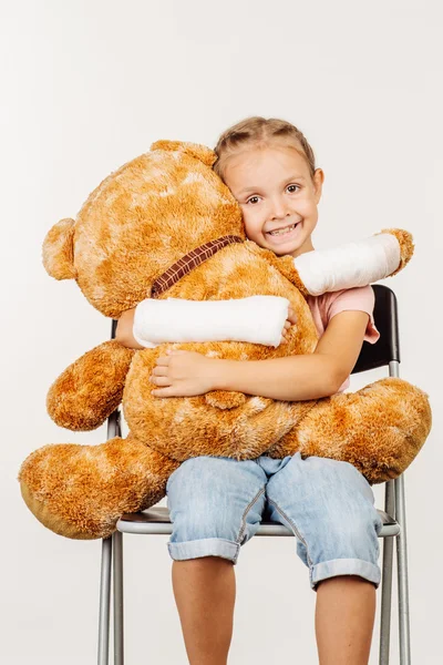 Chica joven con el brazo roto está sonriendo y abrazando a un oso de juguete suave —  Fotos de Stock