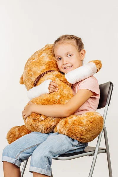 Mladá dívka se zlomenou rukou se usmívá a objímání Plyšová hračka medvěd — Stock fotografie