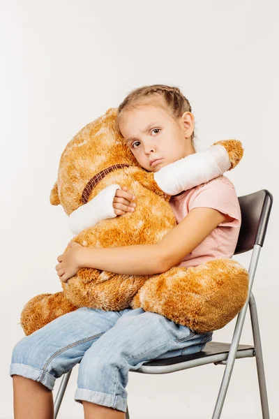 Jovem menina triste com braço quebrado está abraçando um urso de brinquedo macio . — Fotografia de Stock