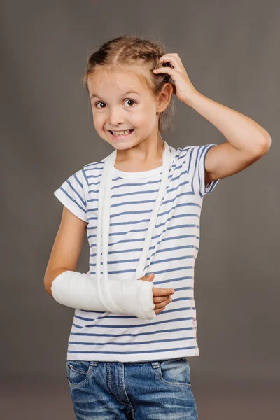 Menina feliz com braço quebrado está de pé sobre o fundo cinza — Fotografia de Stock