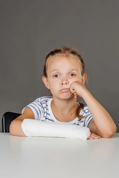 Uttråkad flicka med bruten arm sitter vid bordet. Medicin — Stockfoto