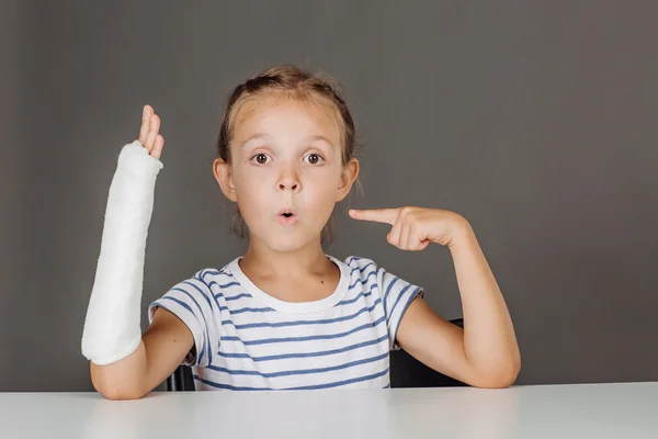 Młoda dziewczyna zaskoczony ze złamaną rękę siedzi przy stole. — Zdjęcie stockowe