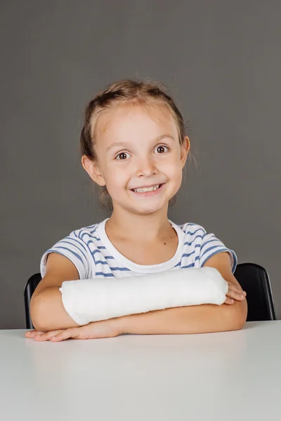 Młoda dziewczyna uśmiechający się ze złamaną rękę siedzi przy stole. Medycyna — Zdjęcie stockowe