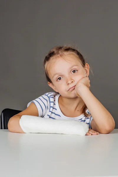 Ledsen flicka med bruten arm sitter vid bordet. Medicin — Stockfoto