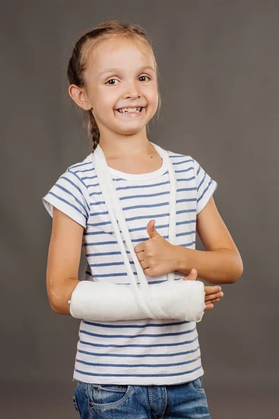 Menina feliz com o braço quebrado está de pé sobre o fundo cinza. M — Fotografia de Stock