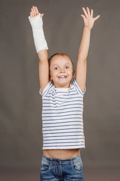 Happy young girl with broken arm is standing on the gray backgro — Stock fotografie