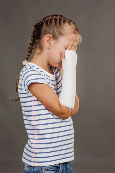 Sad girl with broken arm is standing on the gray background. Med — Stockfoto