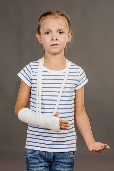 Menina assustada com o braço quebrado está de pé sobre o fundo cinza . — Fotografia de Stock