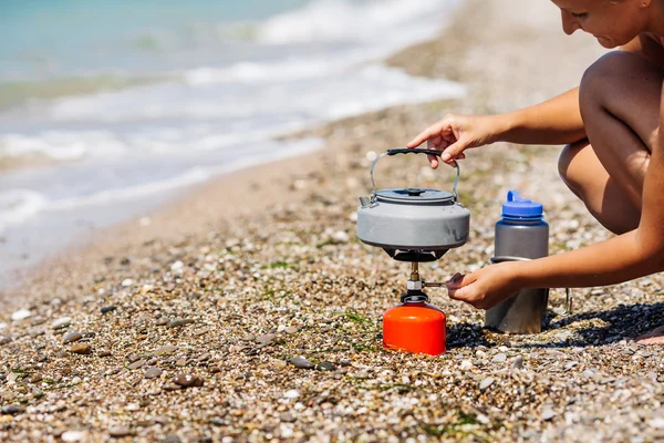 Woman boiling water on camping stove on sea shore.