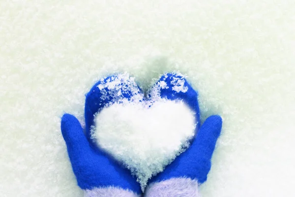 Hands in knitted mittens with a heart made of snow on a winter day. Snowy heart in hands. Human hands in warm blue mittens with a snowy heart on a background of snow. Love winter christmas or romantic — Stock Photo, Image