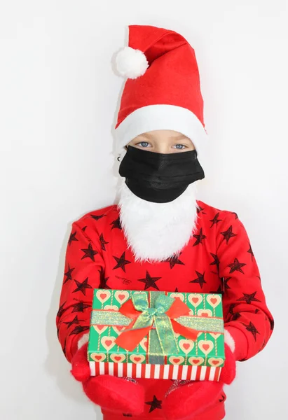 Mały Mikołaj daje prezenty dla kwarantanny na niego maskę od wirusa. czerwony sweter Boże Narodzenie i Nowy Rok prezent. Happy little girl w Santa s kapelusz ma Boże Narodzenie. — Zdjęcie stockowe