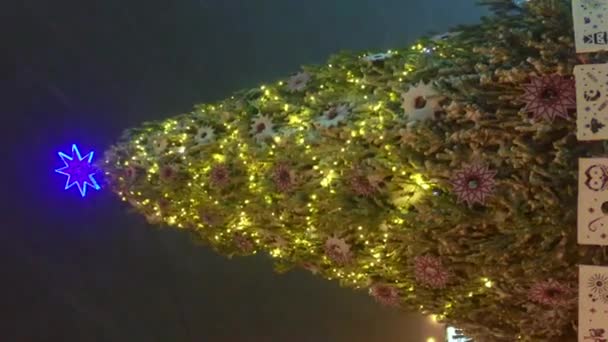 あなたの携帯電話のための垂直ビデオクリスマスイブ、大きな装飾されたクリスマスツリーは正方形に立っていて、ガーランドのカラフルな光でちらつきます。冬の雪の日。都市照明のガーランドビュー — ストック動画