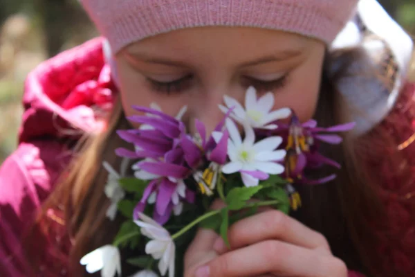 Liten flicka samlar blommor på en äng i skogen. Flicka i rosa. En mörk skog och de första vårblommorna i solen. — Stockfoto