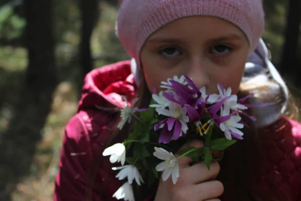 Little girl collects flowers in a meadow in the forest. Girl in pink. A dark forest and the first spring flowers in the sun. — Stock Photo, Image