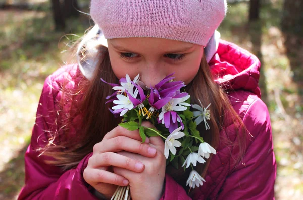 Liten flicka samlar blommor på en äng i skogen. Flicka i rosa. En mörk skog och de första vårblommorna i solen. — Stockfoto