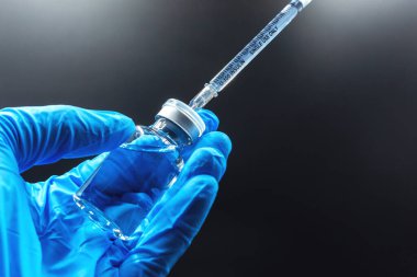 Aşı kavramı, doktorun beyaz aşı şişesi ve siyah arka planda izole edilmiş fotokopi alanı olan mavi eldivenin elinde.