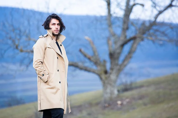 Image of young beautiful man with long hair wearing rain jacket in cold weather. Thinker european man in spring nature.