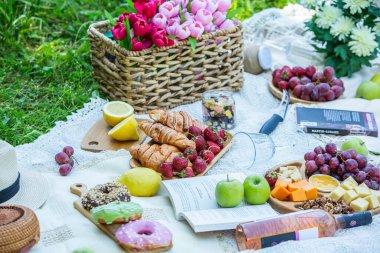 Outdoors picnic in a lush green park with a tasty croissant, fruits, donuts and wine on grass. Summer picnic on the blanket.