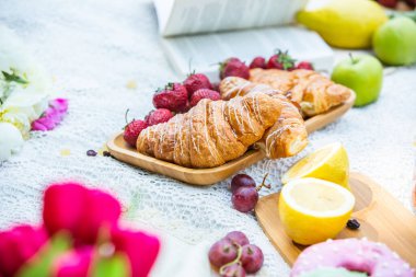 Outdoors picnic in a lush green park with a tasty croissant, fruits, donuts and wine on grass. Summer picnic on the blanket.