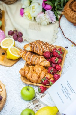 Outdoors picnic in a lush green park with a tasty croissant, fruits, donuts and wine on grass. Summer picnic on the blanket.