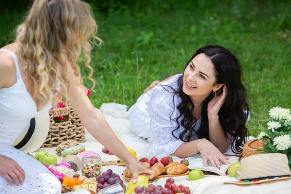 Two girls are resting in park sitting on a picnic blanket. Friends is making picnic outdoor with fruits and wine. Blonde and brunette girl in nature.
