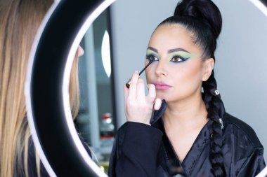 Beautiful European woman getting artistic makeup applied by a professional makeup artist in a modern beauty salon, glamour and elegance concept.