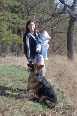 Güneşli bir günde bebeğiyle ve köpeğiyle ormanda yürüyen bir kadın.