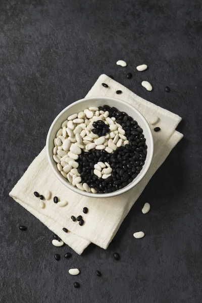 Beans on a dark background. White beans and black beans in a gray bowl on a napkin. Sign Yin Yang. Top view. Vertical position.