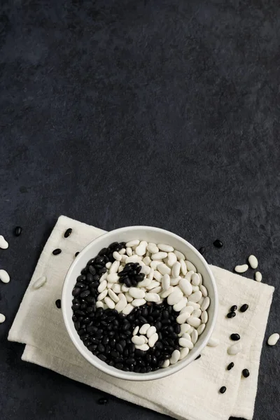 Beans on a dark background. White beans and black beans in a gray bowl on a napkin. Sign Yin Yang. Top view. Vertical position. Copy space