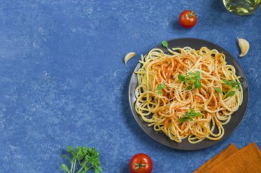 Geleneksel İtalyan spagettisi, siyah yuvarlak tabakta, mavi arka planda. Makarna için malzemeler - kirazlı domates, parmesan, sarımsak, zeytinyağı, fesleğen. Kopyalama alanı olan üst görünüm. Yatay konum.