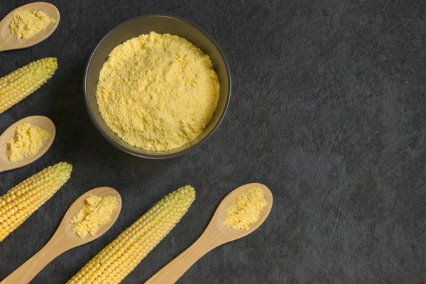 Fresh corn on the cob and corn grits in a bowl and wooden spoons on a dark background. The concept of production from corn grains. Top view. Copy space.