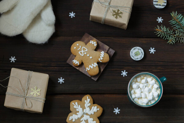 Christmas New Year card.Cup of hot drink with marshmallows, warm white mittens, New Year's gift, gingerbread, snowflakes, candles on a dark wooden background. Festive concept. Top view