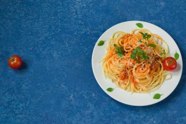 Geleneksel İtalyan spagettisi bolonez beyaz yuvarlak tabakta mavi arka planda. Makarna için malzemeler - kirazlı domates, parmesan, sarımsak, zeytinyağı, fesleğen. Kopyalama alanı olan üst görünüm. Yatay konum