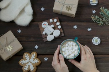 Dişi eller bir fincan sıcak içeceği şekerle birlikte tutarlar. Sıcak beyaz eldivenler, yeni yıl hediyesi, marshmallow kardan adam, zencefilli ekmek, koyu ahşap arka planda mumlar. Yılbaşı kartı.