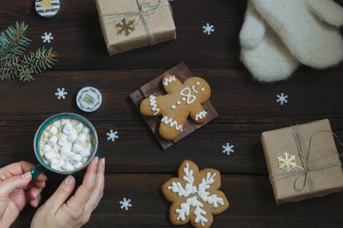 Dişi eller bir fincan sıcak içeceği şekerle birlikte tutarlar. Sıcak beyaz eldivenler, yeni yıl hediyesi, zencefilli çörek, koyu ahşap arka planda mumlar. Yılbaşı kartı. Yukarıdan görüntüle.
