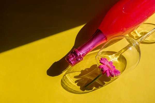 Festive party concept. Hard light hard shadows. Glass and bottle of pink champagne on a yellow background. Sunlight reveals the solid forms of glass and bottle shadows. Flat lay.