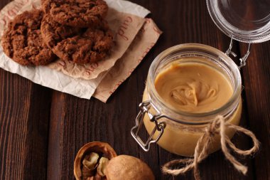 Doğal fıstık ezmeli cam kavanoz, sağlıklı kurabiyeler, kahverengi ahşap arka planda ceviz. Vejetaryen geleneksel kahvaltısı